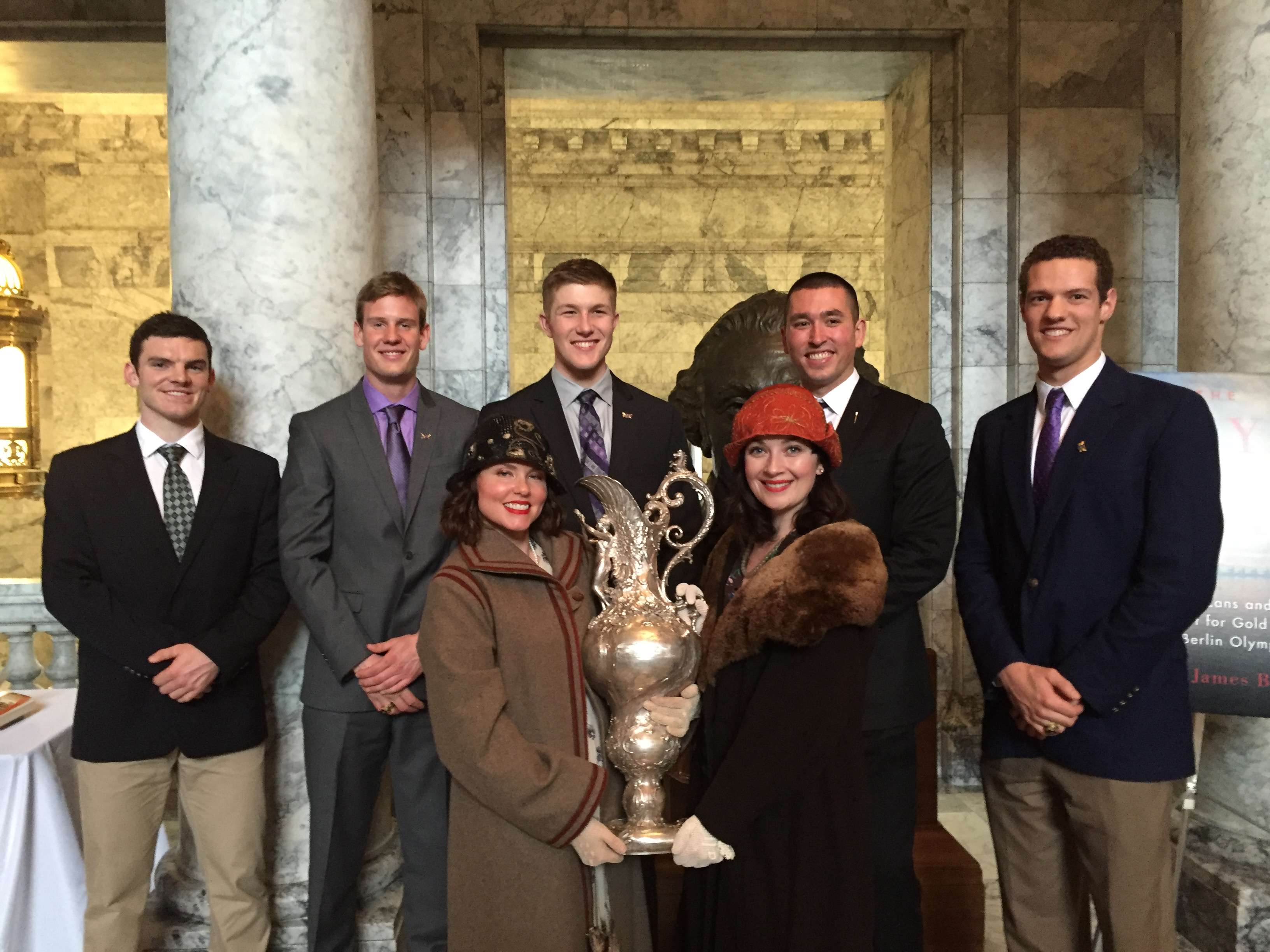 Actors Lisa Coronado and Carollani Sandberg pose in "The Boys in the Boat" costume with members of the UW crew team and their Olympic trophy from the team's historic 1936 victory.